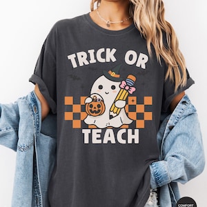 K&ouml;nnte beinhalten: Ein dunkelgraues T-Shirt mit dem Text "Trick or Teach" und einem Cartoon-Geist, der einen Bleistift und einen K&uuml;rbis h&auml;lt. Der Geist tr&auml;gt einen Hexenhut und eine rosafarbene Schleife.