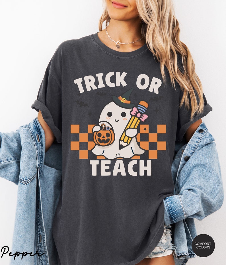 K&ouml;nnte beinhalten: Ein dunkelgraues T-Shirt mit dem Text "Trick or Teach" und einem Cartoon-Geist, der einen Bleistift und einen K&uuml;rbis h&auml;lt. Der Geist tr&auml;gt einen Hexenhut und eine rosafarbene Schleife.