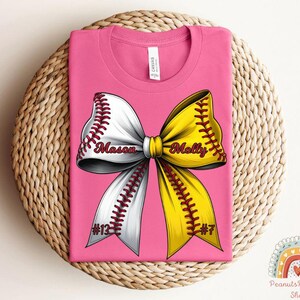 May include: Pink t-shirt with a white and yellow softball bow. The bow has the names "Mason" and "Molly" written on it. The shirt also has the numbers "#12" and "#7" on it.