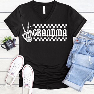 May include: Black v-neck t-shirt with a white graphic of a skeleton hand giving the peace sign and the word "Grandma" in a checkered pattern.