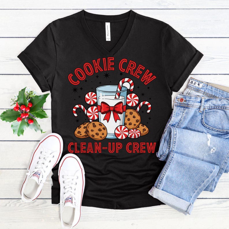 May include: Black V-neck t-shirt with a festive graphic. The design features a glass of milk with a red bow, cookies, candy canes, and the text "COOKIE CREW CLEAN-UP CREW" in red. White sneakers and blue jeans are also visible.