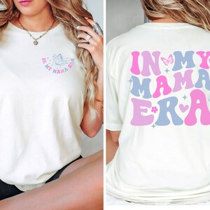 May include: White t-shirt with a graphic on the front that says "In My Mama Era" with a butterfly and a heart. The back of the shirt has a larger graphic that says "In My Mama Era" with butterflies, hearts, and stars in pink and blue.