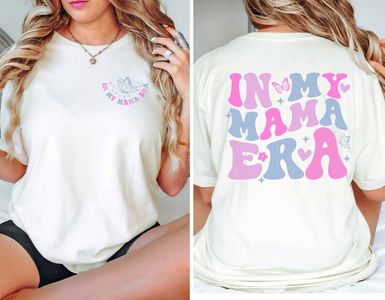 May include: White t-shirt with a graphic on the front that says "In My Mama Era" with a butterfly and a heart. The back of the shirt has a larger graphic that says "In My Mama Era" with butterflies, hearts, and stars in pink and blue.