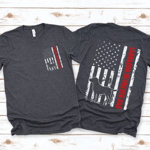 May include: Two dark heather grey t-shirts with a distressed American flag design. One shirt has a silhouette of a Labrador Retriever and the text "Labrador Retriever Dad" on the back. The other shirt has a silhouette of a Labrador Retriever on the front.