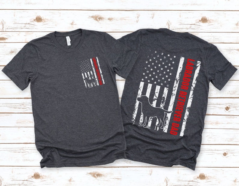 May include: Two dark heather grey t-shirts with a distressed American flag design. One shirt has a silhouette of a Labrador Retriever and the text "Labrador Retriever Dad" on the back. The other shirt has a silhouette of a Labrador Retriever on the front.