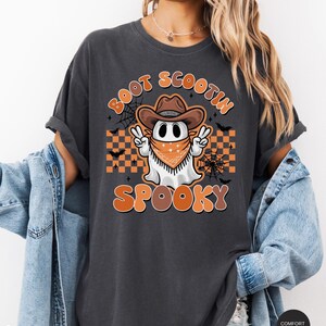 May include: A dark gray t-shirt with a cartoon ghost wearing a cowboy hat and bandana. The ghost is holding up two fingers in a peace sign. The text "Boot Scootin' Spooky" is printed on the shirt in orange and black.