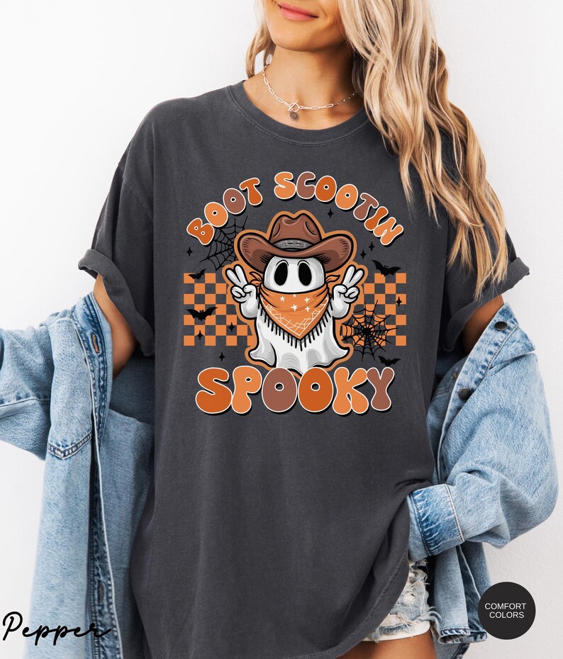 May include: A dark gray t-shirt with a cartoon ghost wearing a cowboy hat and bandana. The ghost is holding up two fingers in a peace sign. The text "Boot Scootin' Spooky" is printed on the shirt in orange and black.
