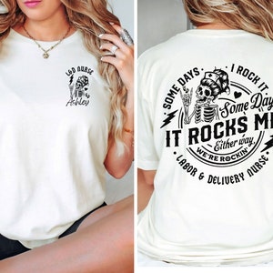 K&ouml;nnte beinhalten: Wei&szlig;es T-Shirt mit einer schwarzen Grafik eines Skeletts, das eine Krankenschwesterm&uuml;tze tr&auml;gt und ein Mikrofon h&auml;lt. Der Text auf dem Shirt lautet "Some days I rock it, Some days it rocks me, Either way, we're rockin', Labor & Delivery Nurse".