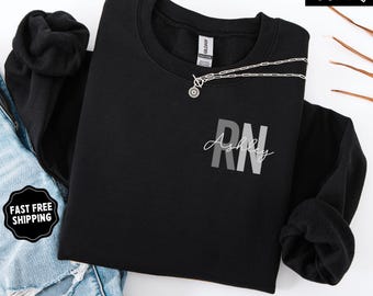 May include: Black crewneck sweatshirt with the letters "RN" in gray and the name "Ashley" in script. A silver necklace is draped across the front. The words "Super Soft" and "Black" are also visible.