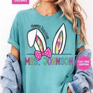 May include: A turquoise blue t-shirt with a white bunny rabbit ear design. The text "Hoppy Teacher" is written in black on the shirt. The text "MRS. JOHNSON" is written in rainbow colors below the bunny ears.