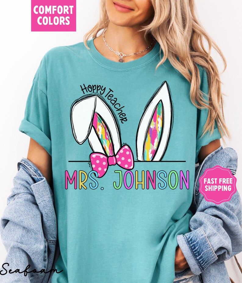 May include: A turquoise blue t-shirt with a white bunny rabbit ear design. The text "Hoppy Teacher" is written in black on the shirt. The text "MRS. JOHNSON" is written in rainbow colors below the bunny ears.