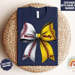 May include: Navy blue t-shirt with a white and yellow softball bow. The bow has red stitching and the names "Mason" and "Molly" with numbers "#13" and "#7" on each side.