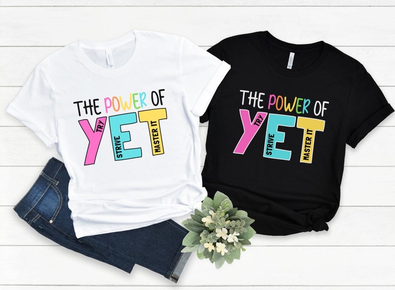 May include: Two t-shirts, one white and one black, with the text "THE POWER OF YET" in colourful block letters. The white shirt is paired with blue jeans. The text includes the words "TRY", "STRIVE", and "MASTER IT".