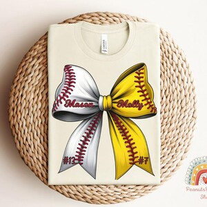 May include: A beige t-shirt with a softball bow design. The bow is white and yellow with red stitching. The words "Mason #12" and "Molly #7" are written on the bow.