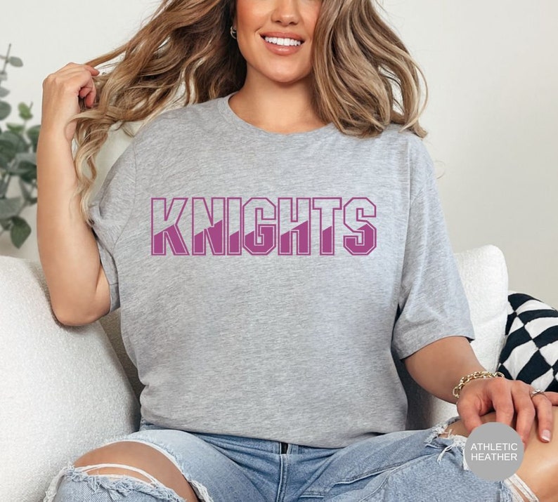 May include: A heather grey t-shirt with the word "KNIGHTS" printed in pink on the front. The shirt is a relaxed fit and has a crew neck.