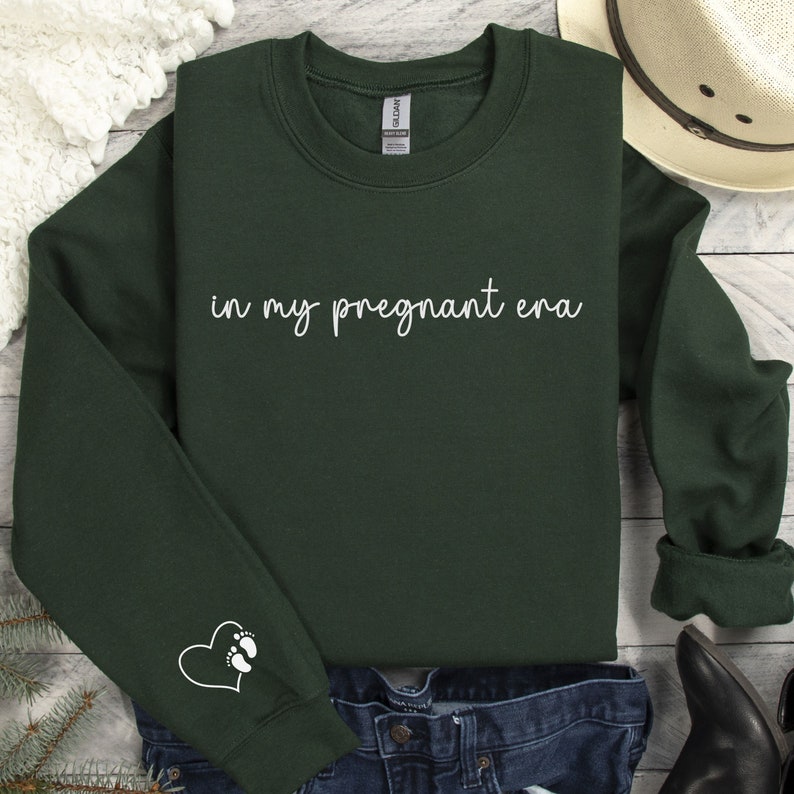 K&ouml;nnte beinhalten: Ein dunkelgr&uuml;ner Pullover mit dem Text "in my pregnant era" in wei&szlig;en Buchstaben. Der linke &Auml;rmel hat ein wei&szlig;es Herz mit einem Fu&szlig;abdruck darin.