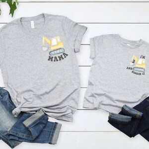 Peut inclure: Deux t-shirts gris avec un motif de pelle m&eacute;canique jaune. L'un dit "MAMA" et l'autre "I'M 3 AND DIGGIN IT". Les deux t-shirts sont associ&eacute;s &agrave; des jeans bleus. Les t-shirts sont sur une surface en bois blanc.