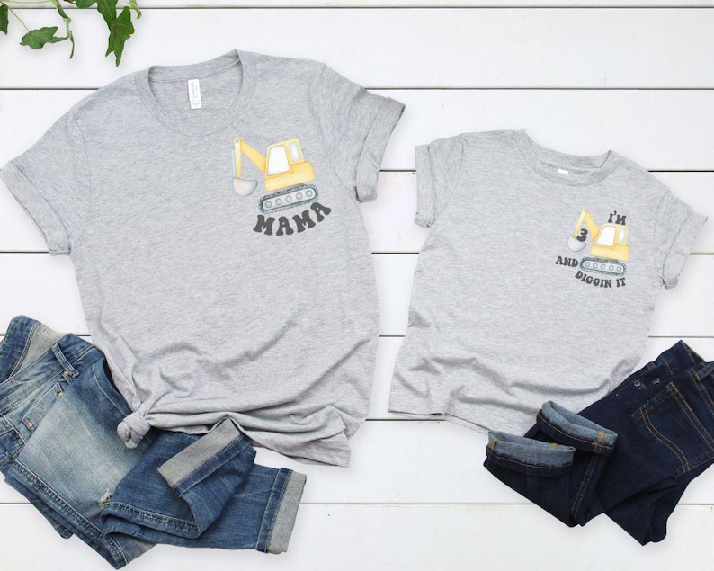 Peut inclure: Deux t-shirts gris avec un motif de pelle m&eacute;canique jaune. L'un dit "MAMA" et l'autre "I'M 3 AND DIGGIN IT". Les deux t-shirts sont associ&eacute;s &agrave; des jeans bleus. Les t-shirts sont sur une surface en bois blanc.