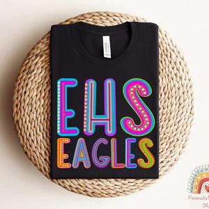 Puede incluir: Camiseta negra con el texto "EHS Eagles" en letras de colores y contorneadas. Las letras est&aacute;n decoradas con peque&ntilde;os puntos de colores.