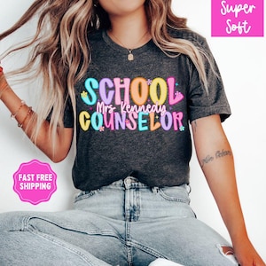 May include: Dark gray t-shirt with the words "SCHOOL Mrs. Kennedy COUNSELOR" in colorful bubble letters. The shirt has a crew neck and short sleeves. The words "Super Soft" are in a pink box in the upper right corner.