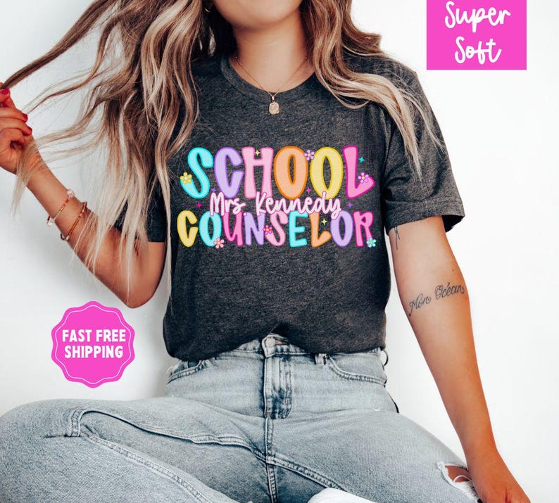 May include: Dark gray t-shirt with the words "SCHOOL Mrs. Kennedy COUNSELOR" in colorful bubble letters. The shirt has a crew neck and short sleeves. The words "Super Soft" are in a pink box in the upper right corner.