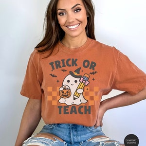 K&ouml;nnte beinhalten: Ein orangefarbenes T-Shirt mit dem Text "Trick or Teach" in Schwarz. Ein Cartoon-Geist mit einer Hexenm&uuml;tze und einem Bleistift und einem K&uuml;rbis-Eimer in der Hand befindet sich in der Mitte des Shirts.