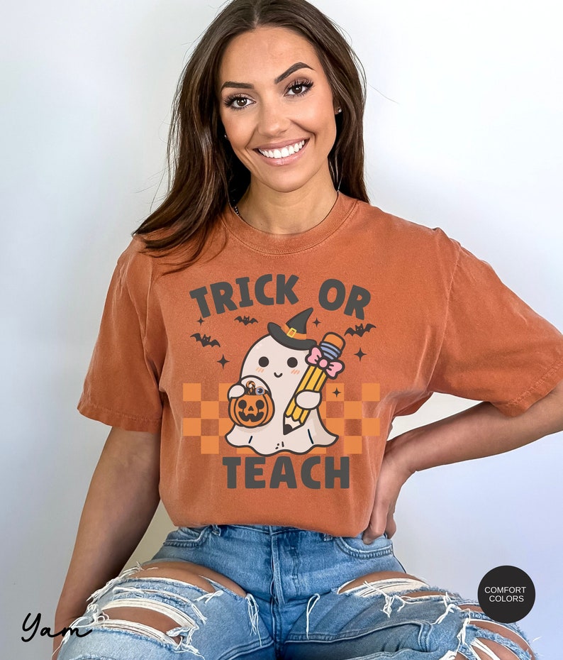 K&ouml;nnte beinhalten: Ein orangefarbenes T-Shirt mit dem Text "Trick or Teach" in Schwarz. Ein Cartoon-Geist mit einer Hexenm&uuml;tze und einem Bleistift und einem K&uuml;rbis-Eimer in der Hand befindet sich in der Mitte des Shirts.