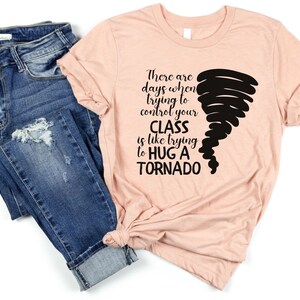 May include: A peach-colored t-shirt with black text that reads "There are days when trying to control your CLASS is like trying to HUG A TORNADO". A black tornado graphic is on the right side of the shirt.
