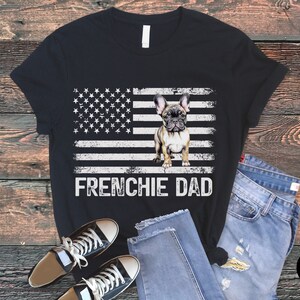 May include: Black t-shirt with a distressed American flag design and a French Bulldog standing in front of it. The text "Frenchie Dad" is printed below the flag.