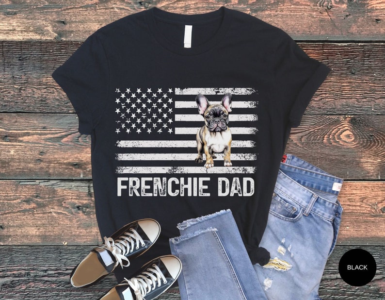 May include: Black t-shirt with a distressed American flag design and a French Bulldog standing in front of it. The text "Frenchie Dad" is printed below the flag.