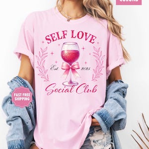 May include: Pink t-shirt with the text "Self Love Social Club" and a glass of wine with a pink bow. The text "Est. 2023" is below the wine glass.