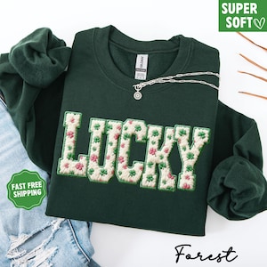 Pu&ograve; includere: Una felpa verde scuro con la scritta "LUCKY" ricamata in bianco con un motivo floreale di trifogli rosa e verdi. Una collana d'argento con un ciondolo &egrave; sulla felpa. Il testo "SUPER SOFT" e "FAST FREE SHIPPING" sono visibili.