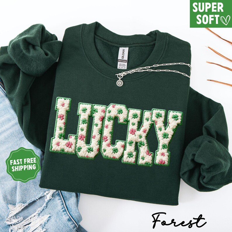 Pu&ograve; includere: Una felpa verde scuro con la scritta "LUCKY" ricamata in bianco con un motivo floreale di trifogli rosa e verdi. Una collana d'argento con un ciondolo &egrave; sulla felpa. Il testo "SUPER SOFT" e "FAST FREE SHIPPING" sono visibili.