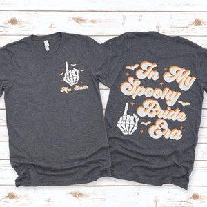 Puede incluir: Dos camisetas de color gris oscuro con un gr&aacute;fico de mano de esqueleto y el texto "In My Spooky Bride Era". La mano de esqueleto lleva un anillo en su dedo &iacute;ndice.