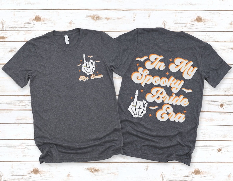 Puede incluir: Dos camisetas de color gris oscuro con un gr&aacute;fico de mano de esqueleto y el texto "In My Spooky Bride Era". La mano de esqueleto lleva un anillo en su dedo &iacute;ndice.