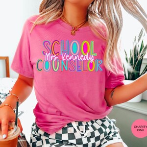 May include: Pink t-shirt with the text "SCHOOL COUNSELOR" in colorful letters. The text "Mrs. Kennedy" is written in black letters below the main text.