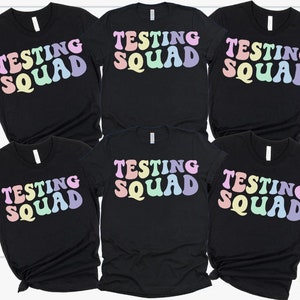May include: Six black t-shirts with the text "Testing Squad" in rainbow colors. The text is in a retro font style.