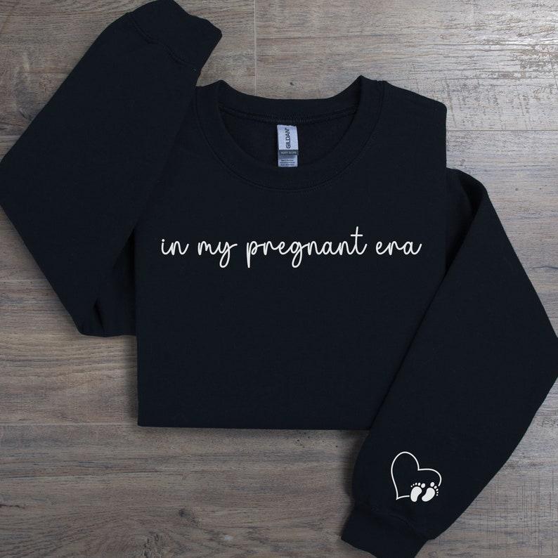 K&ouml;nnte beinhalten: Schwarzer Pullover mit dem Text "in my pregnant era" in wei&szlig;er kursivschrift. Ein wei&szlig;es Herz mit Fu&szlig;abdr&uuml;cken befindet sich auf dem linken &Auml;rmel.