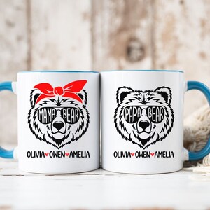Op de afbeelding: Twee witte keramische mokken met blauwe randen. De mok aan de linkerkant heeft een zwart-witte illustratie van een beer met een zonnebril en een rode bandana met de tekst "Mama Bear" en de namen "Olivia Owen Amelia" eronder. De mok aan de rechterkant heeft een zwart-witte illustratie van een beer met een zonnebril met de tekst "Papa Bear" en de namen "Olivia Owen Amelia" eronder.