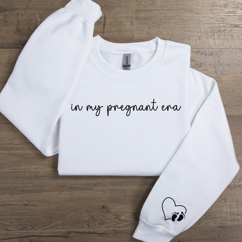 K&ouml;nnte beinhalten: Wei&szlig;er Pullover mit dem Text "in my pregnant era" in schwarzer Schrift. Ein schwarzes Herz mit zwei Fu&szlig;abdr&uuml;cken befindet sich auf dem linken &Auml;rmel.