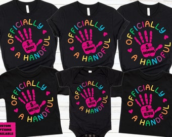 Könnte beinhalten: Fünf schwarze T-Shirts mit einem bunten Handabdruck-Design und dem Text "Officially a Handful" mit verschiedenen Titeln für jedes Shirt: "Daddy", "Mommy", "Grandma", "Brother" und "Sister".