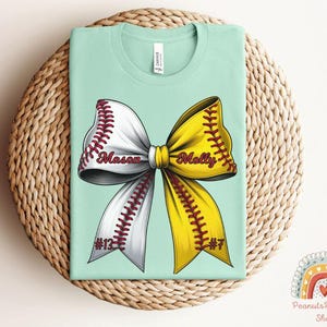 May include: A light blue t-shirt with a softball bow design. The bow is yellow and white with red stitching. The words "Mason" and "Molly" are written on the bow. The numbers "#13" and "#7" are on the bottom of the bow.