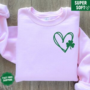 May include: A pink sweatshirt featuring a green heart and shamrock design. The heart is drawn in a simple, hand-drawn style.