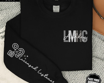 May include: Black sweatshirt with the text "LMHC Ashley" in white and gray. The left sleeve features a graphic of an ear, speech bubble, and stars, with the words "Licensed Listener" in white. The upper left corner has "SUPER SOFT" text.