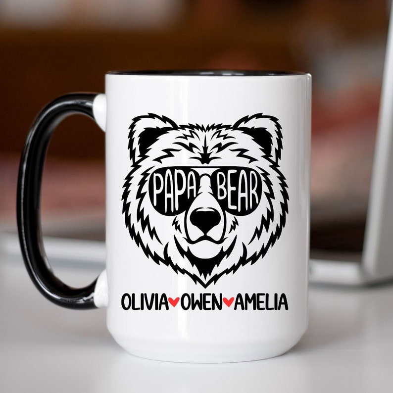 Op de afbeelding: Een witte keramische mok met een zwarte rand en handvat. De mok heeft een zwart-witte illustratie van een beer met een zonnebril met de tekst "Papa Bear" boven het hoofd van de beer. Onder de beer staat de tekst "Olivia Owen Amelia" gedrukt met rode hartjes tussen de namen.