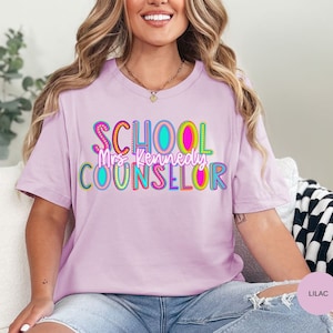 May include: Lilac t-shirt with the text "SCHOOL COUNSELOR" in colorful letters with a rainbow design. The text "Mrs. Kennedy" is written in a smaller font below the main text.