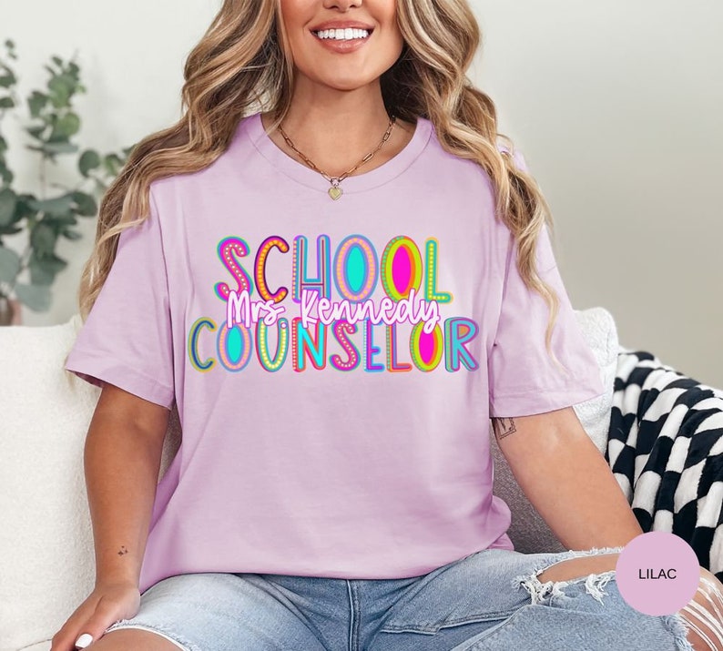 May include: Lilac t-shirt with the text "SCHOOL COUNSELOR" in colorful letters with a rainbow design. The text "Mrs. Kennedy" is written in a smaller font below the main text.