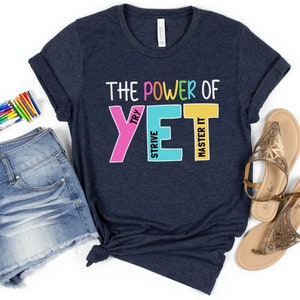 May include: A navy blue t-shirt with the phrase "THE POWER OF YET" in colourful block letters. The shirt is styled with denim shorts, sandals, and a set of coloured pencils, suggesting a motivational or back-to-school theme.