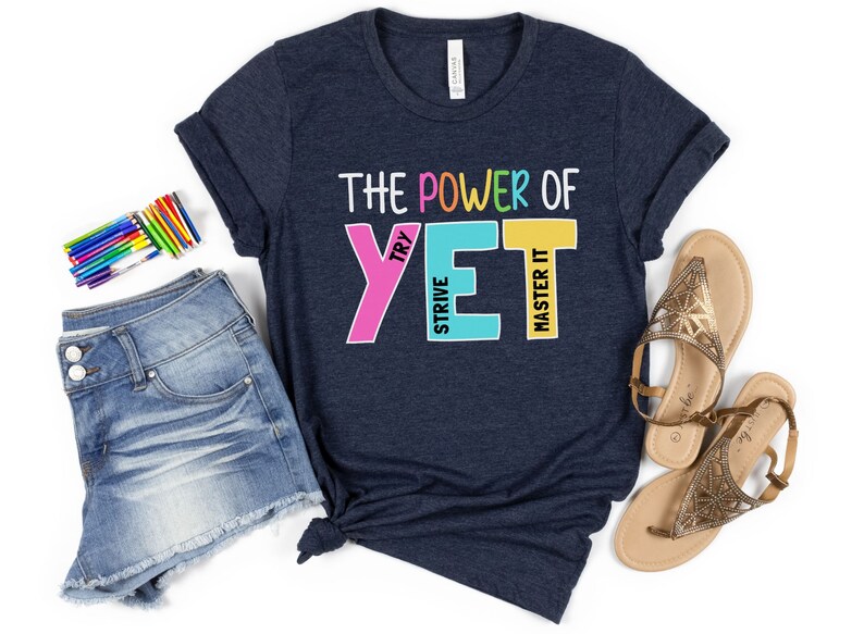 May include: A navy blue t-shirt with the phrase "THE POWER OF YET" in colourful block letters. The shirt is styled with denim shorts, sandals, and a set of coloured pencils, suggesting a motivational or back-to-school theme.