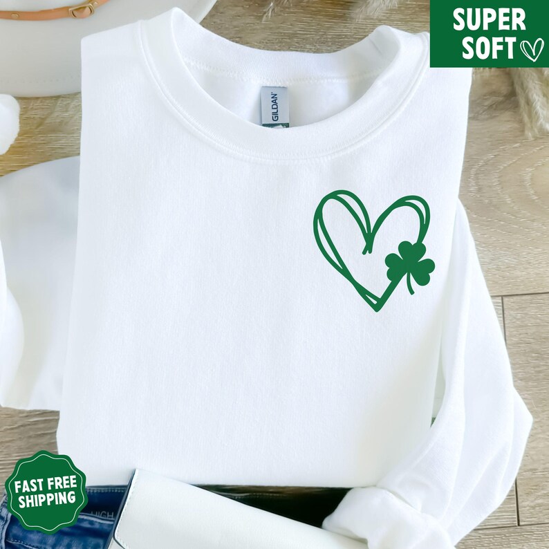 May include: White sweatshirt featuring a green heart and shamrock design. The heart is drawn in a simple, hand-drawn style.